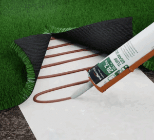 A hand applies Seam Adhesive 1 Gallon, Green—covering 40 linear ft—onto white seam tape between two artificial grass pieces, preparing to bond the turf sections.